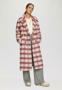 Dubbelzijdige wollen jas met een roze en crème geruit patroon, grote knopen en een reverskraag, gecombineerd met grijze wijde broekspijpen.