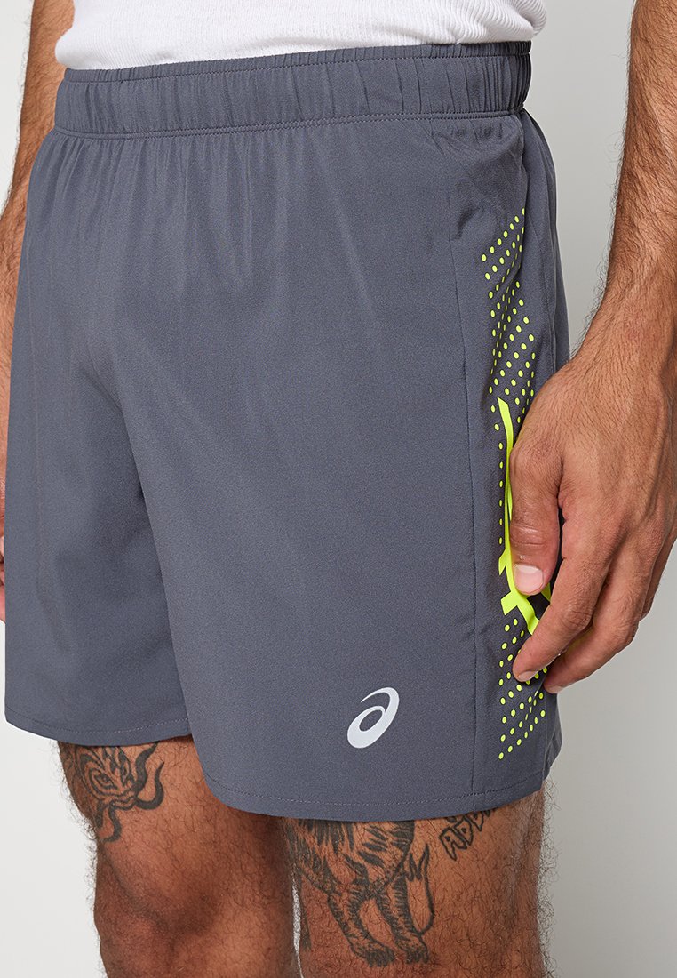 Grijze sportshorts met een neongele zijontwerp en witte logo, gedragen door een getatoeëerde mannelijke model met zijn hand op zijn dij.