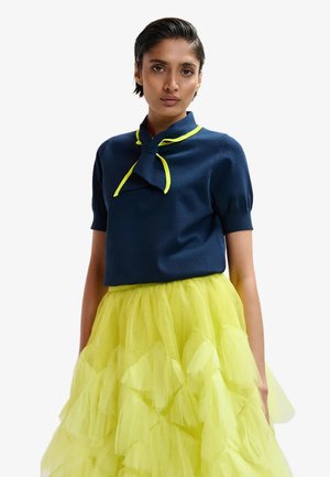 Donna con capelli corti e scuri che indossa una maglietta a maniche corte blu navy con un fiocco bordato di giallo e una gonna a strati di tulle giallo acceso.