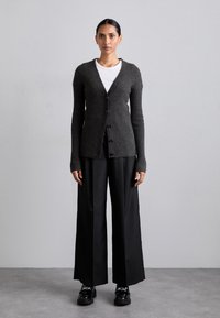 Cardigan gris foncé côtelé avec un col en V, des manches longues et des boutons sur le devant. Porté sur une chemise blanche et associé à un pantalon noir à jambes larges.