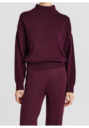 Femme portant un pull en maille violet foncé assorti avec col montant et un pantalon ample en maille sur un fond gris uni.
