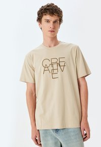 Beige bomulds-t-shirt med "CREATIVE" grafik trykt lodret i brun. Rund halsudskæring og korte ærmer; parret med lyseblå jeans.