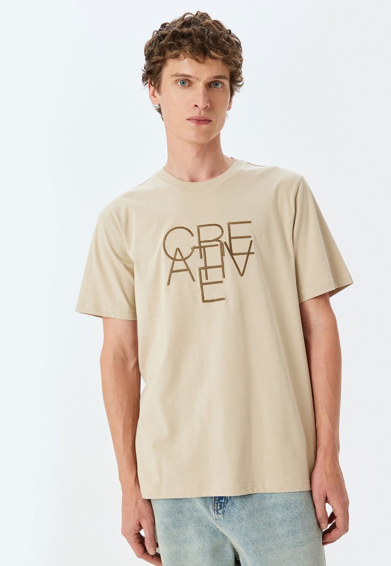 Beige bomulds-t-shirt med "CREATIVE" grafik trykt lodret i brun. Rund halsudskæring og korte ærmer; parret med lyseblå jeans.