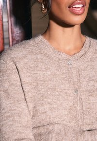 Beżowy sweter na guziki z okrągłym dekoltem, z małymi guzikami i miękką teksturą. Ciekawe detale to subtelny melanżowy wzór.