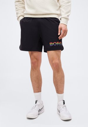 Άνδρας φοράει μαύρα σορτς με πολύχρωμο λογότυπο "BORG" στο αριστερό πόδι, λευκά αθλητικά παπούτσια, λευκές κάλτσες και μπεζ φούτερ.