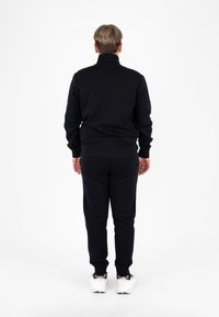 Tuta sportiva nera composta da una giacca con zip e pantaloni abbinati, realizzata in un tessuto morbido; presenta un colletto alto e polsini aderenti.