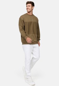 Khakifarbenes Sweatshirt mit einem horizontalen Farbblockdesign, gerippten Bündchen und einem kleinen Logopatch; kombiniert mit weißen Hosen und Sneakers.