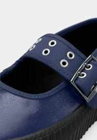 Donkerblauwe Mary Jane-schoen met een gestructureerde band die is voorzien van zilveren oogjes en een gesp, zwarte ribbelzool en een gladde, glanzende afwerking.