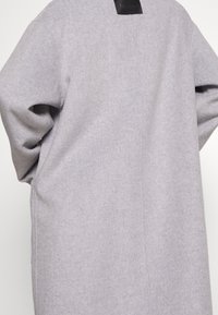 Personne portant un manteau ample en laine gris clair à manches longues, vue de dos sur un fond uni.