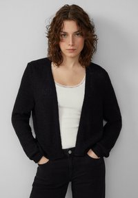 Schwarze Strickjacke mit offenem Vorderteil, gerippter Textur und langen Ärmeln. Getragen über einem weißen Tanktop mit Spitzenrand. Kombiniert mit schwarzen Hosen.