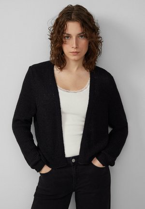 Schwarze Strickjacke mit offenem Vorderteil, gerippter Textur und langen Ärmeln. Getragen über einem weißen Tanktop mit Spitzenrand. Kombiniert mit schwarzen Hosen.