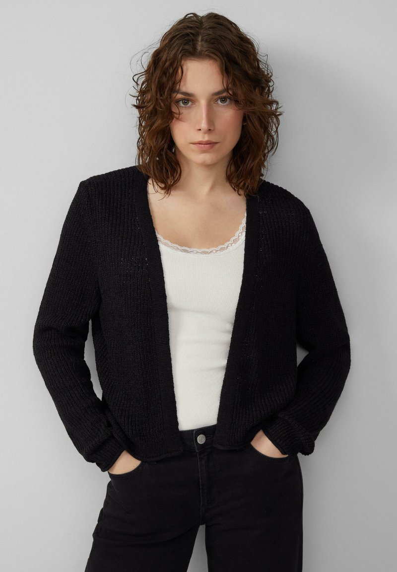 Schwarze Strickjacke mit offenem Vorderteil, gerippter Textur und langen Ärmeln. Getragen über einem weißen Tanktop mit Spitzenrand. Kombiniert mit schwarzen Hosen.