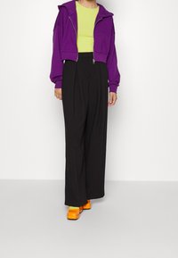 Felpa cropped viola con zip, top a coste verde lime, pantaloni neri a gamba larga e scarpe platform arancioni vivaci. Texture morbide e colori audaci.