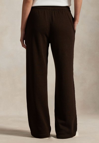 Persona che indossa pantaloni larghi marrone scuro con elastico in vita, mostrato di spalle su uno sfondo neutro e semplice.