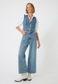 Chaleco de denim con botones, combinado con jeans de pierna ancha y cintura alta. Ambos en lavado azul claro. Usado con botas de cowboy blancas.