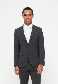 Completo grigio su misura con una vestibilità slim. Caratterizzato da due bottoni, revers a punta e tasche laterali. Indossato sopra una camicia bianca con bottoni.