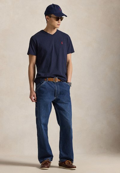 Teget majica s V-izrezom, plave traperice, smeđi remen, smeđe brodske cipele i teget bejzbol kapa. Dodaci uključuju sunčane naočale.