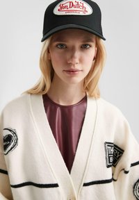 Blondynka w czapce Von Dutch w czerni, w kremowym kardiganie z czarnymi łatami i bordowej bluzce, zwrócona twarzą do kamery z neutralnym wyrazem twarzy.