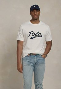 Balta kokvilnas T-kreklu ar tumši zila "Polo" uzdruku un "Ralph Lauren" tekstu, apvienots ar gaiši ziliem džinsiem un tumši zilu beisbola cepuri.