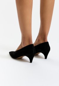 Černé semišové lodičky na vysokém podpatku s špičatou špičkou, nízkým kuželovým podpatkem a elegantním designem. Bez viditelných ozdob nebo dekorací.