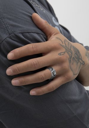 Anillo de plata con patrón grabado, llevado en una mano que reposa sobre una tela gris, destacando la textura suave y el diseño circular.