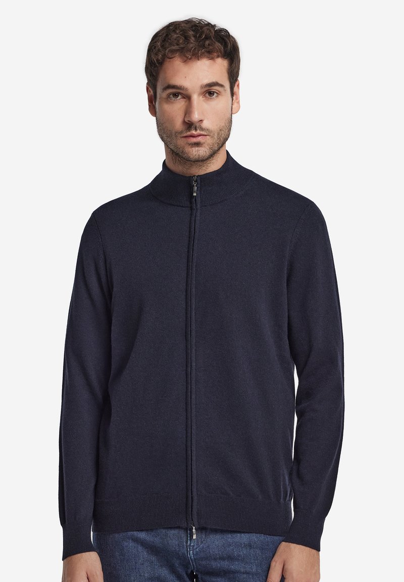 Jonge man draagt een donkerblauwe sweater met ritssluiting en opstaande kraag en een blauwe spijkerbroek, kijkt recht vooruit met een neutrale gezichtsuitdrukking.