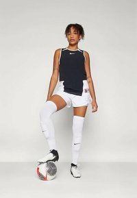 Nike sportsantrekk med en svart ermeløs topp og hvite shorts, kombinert med hvite knelange sokker og svarte fotballsko. En fotball ligger på bakken.