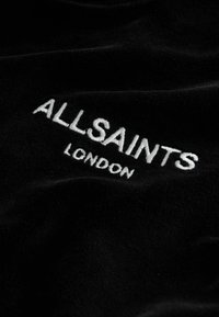 Juodas vilnos audinys su balta siuvinėta fraze "ALLSAINTS" viršuje ir "LONDON" apačioje, akcentuojantis švelnią tekstūrą ir išskirtinį logotipo dizainą.