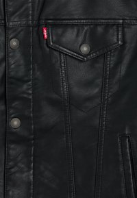 Veste en cuir noir avec des boutons-pression et une poche poitrine. Surface texturée avec des coutures visibles et une petite étiquette de logo.