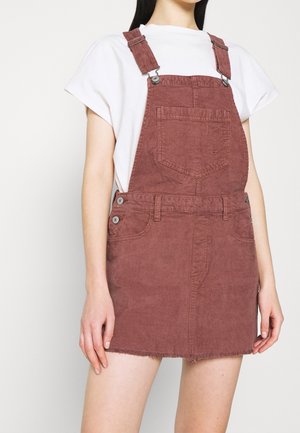 Cordkleid aus Cord in staubigem Rosa mit verstellbaren Trägern, aufgesetzten Taschen vorne und ausgefranster Saum. Über einem weißen T-Shirt getragen. Keine weiteren Details.