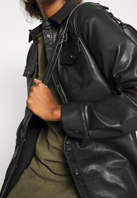 Camisa chaqueta de cuero negro con cierre a presión, que cuenta con dos bolsillos en el pecho y una textura suave. Llevada sobre un suéter verde.