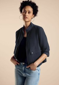 Marineblaue, mit Reißverschluss versehenes Bomberjacke mit Stehkragen, seitlichen Reißverschlusstaschen und kurzen Ärmeln, kombiniert mit hellblauen, hoch taillierten Jeans.