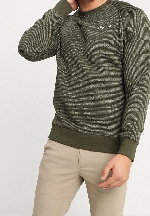 Homme portant un pull texturé vert olive avec le texte « Original » sur la poitrine et un pantalon beige, posant avec une main près du cou.