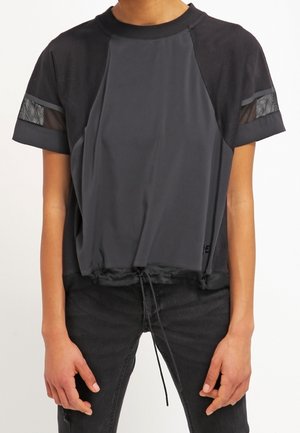 Osoba ubrana w luźną czarno-grafitową koszulkę z krótkim rękawem, z siateczkowatym detalem na rękawach i sznurkiem u dołu.