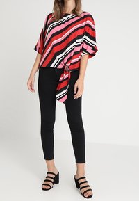 Blusa a righe rossa, nera e rosa con maniche larghe e dettaglio nodo anteriore, abbinata a pantaloni skinny neri e sandali con tacco neri.