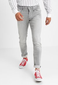 Uomo che indossa jeans grigi chiari arrotolati, sneakers rosse e una camicia bianca con strisce verticali nere, che cammina in avanti su uno sfondo bianco.