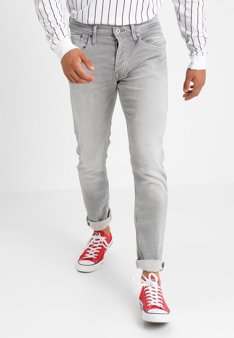 Uomo che indossa jeans grigi chiari arrotolati, sneakers rosse e una camicia bianca con strisce verticali nere, che cammina in avanti su uno sfondo bianco.