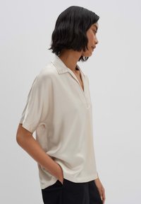 Kurzärmlige, kragenlose Bluse in hellbeige, aus glattem Stoff mit lockerer Passform und einem dezenten Knopfverschluss am Hals.