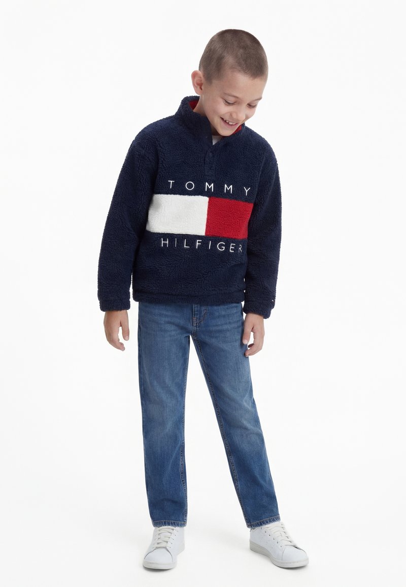 Dreng iført en marineblå Tommy Hilfiger-fleece med hvidt og rødt blokdesign, blå jeans og hvide sneakers, som kigger ned og smiler.