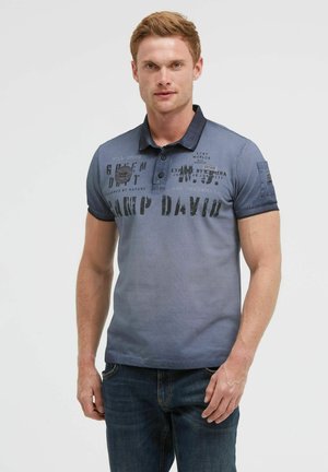 Young man with short light brown hair wearing a blue Camp David polo shirt and dark jeans standing against a plain background.