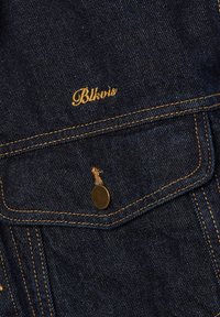 Dunkelblaue Jeansjackentasche mit orangefarbenen Nähten, einem Metallknopf und dem gelb bestickten "Bbkavis"-Logo über der Klappe.