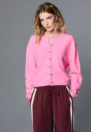 Hölgy hullámos szőke hajjal, élénk rózsaszín gombos kardigánt és bordó színű, fehér oldalsávos melegítőnadrágot visel, szürke háttér előtt áll.