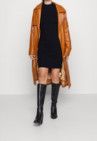 Cappotto lungo in pelle marrone sopra un vestito in maglia nera a coste, abbinato a stivali al ginocchio neri con tacco a blocco. Texture morbide e silhouette moderna.