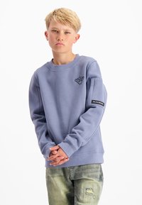 Blonde jongen met een serieuze uitdrukking, gekleed in een blauwe sweatshirt en distressed groene jeans, staande voor een witte achtergrond.