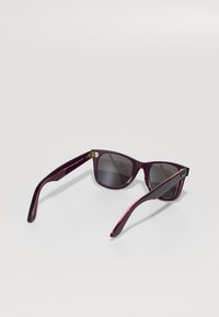 Na bijelom pozadinskom okviru, ljubičaste Ray-Ban sunčane naočale s tamnim lećama, preklopljenim ručkama i vidljivim logotipom marke na desnoj ručki.