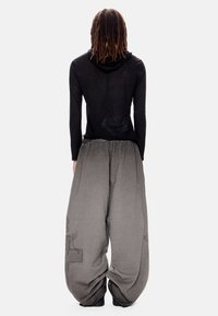 Άτομο με μακριές πλεξούδες που φοράει μαύρο τοπ με μακριά μανίκια και χαλαρό, φαρδύ γκρι παντελόνι με διακριτικά σχέδια, στέκεται με την πλάτη προς το μέρος μας.