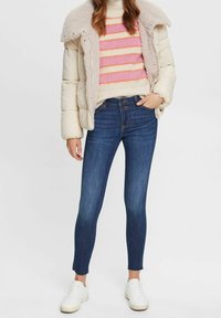 Chaqueta acolchada en crema con cuello de borrego, suéter a rayas en rosa y naranja, jeans ajustados azul oscuro y zapatillas blancas.