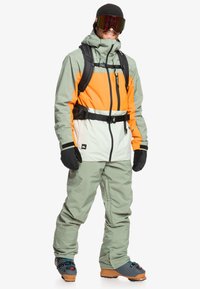 Skikleding in gedempt groen, oranje en wit. Heeft een capuchon, verstelbare taille, handschoenen en een skibril. Textuurstof, functioneel ontwerp.