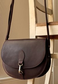 Brown leather crossbody bag with a gold clasp hanging on the back of a worn white wooden chair against a beige wall.