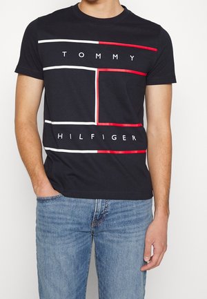 Homme portant un T-shirt noir Tommy Hilfiger avec des lignes rectangulaires blanches et rouges et un jean bleu, debout avec une main dans la poche.